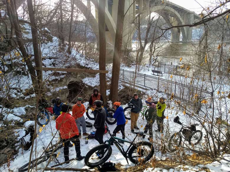 A group of cyclists and their bikes gather under a river bridge in the woods on a patch of snow