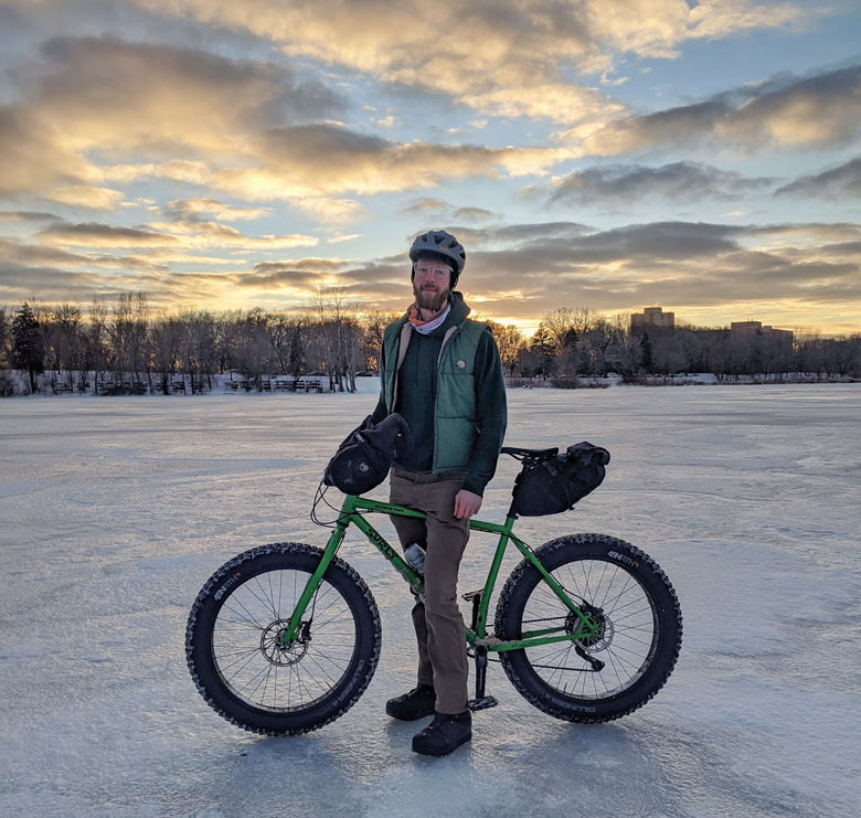 Cyclist stands on frozen lake with Surly Ice Cream Truck fat bike with seat bag, water bottle, and trees in the distance