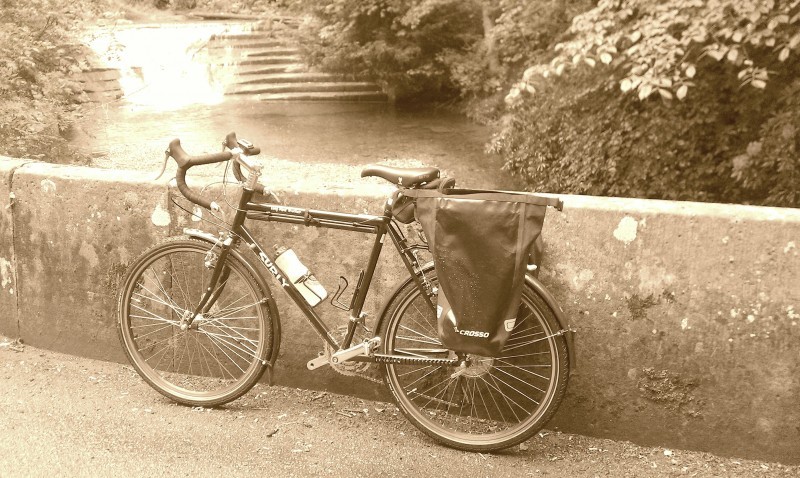 Left side view of a Surly Long Haul Trucker bike, leaning on a cement wall with a waterfall behind it - black & white