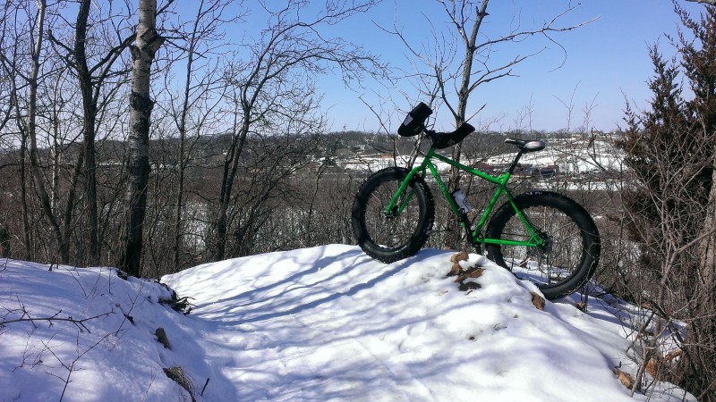 Left side view of a green Surly fat bike, parked on a snowbank on the side of a snow covered trail in the woods