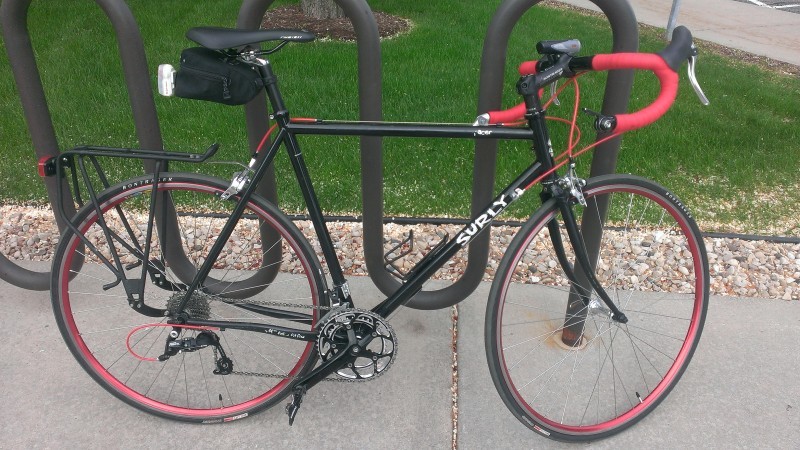 Right side view of a black Surly Pacer bike with red handlebar, leaning along a bike rack on a sidewalk