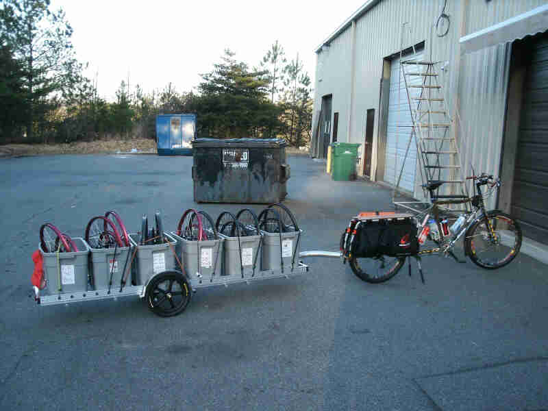 Right side view of a Surly Big Dummy bike with a trailer of bins, full of bike rims, on an outdoor warehouse lot