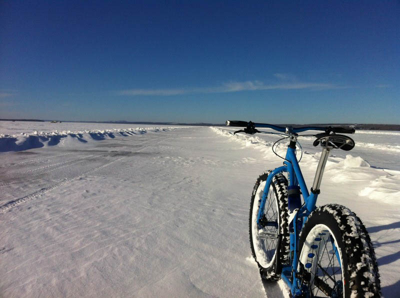 Rear view of a Surly fat bike, blue, in snow, facing down a wide roadway on a frozen, snowy lake