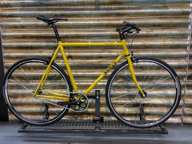 Right profile of a Surly Wednesday bike, yellow, parked in front of a steel wall 
