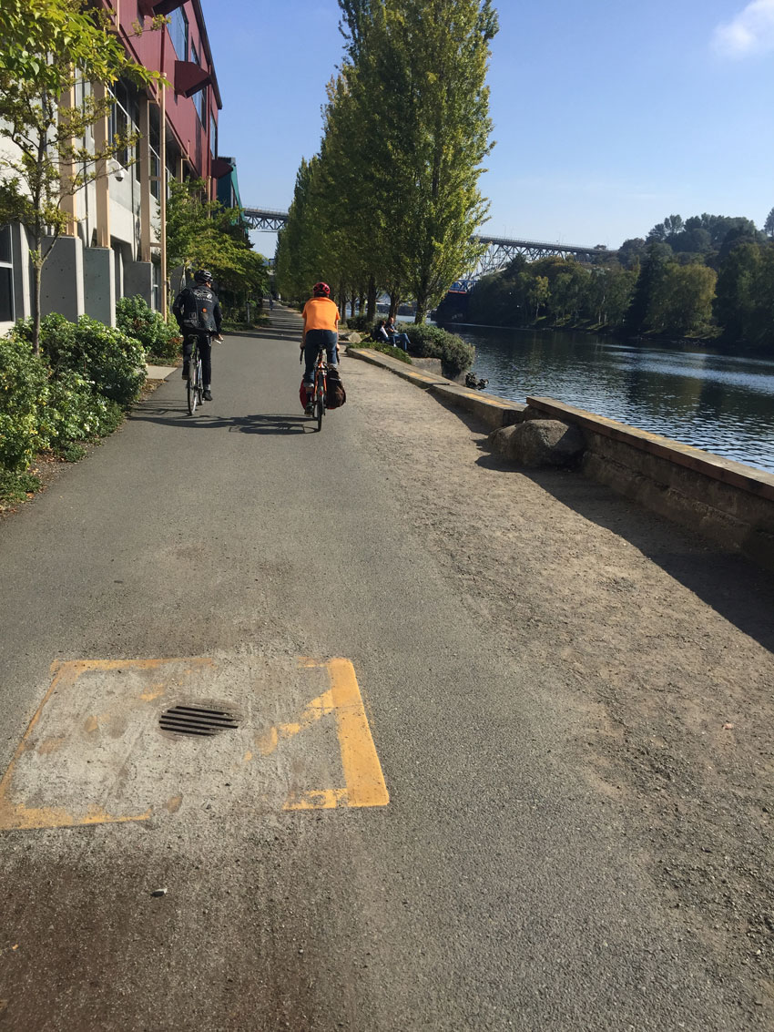 Rear view of two cyclists, riding their bikes down a road along a river on one side, with a building on their other side