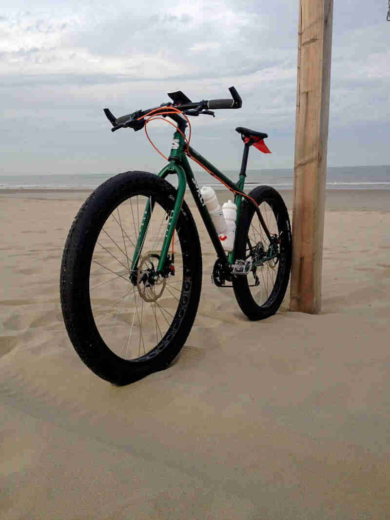 Front left side view of a Surly Krampus bike, green, on a sandy beach, with the ocean in the background