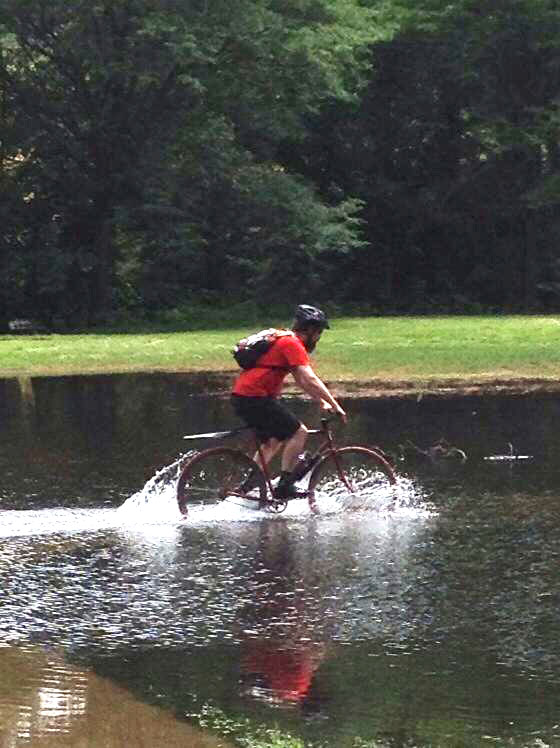 Right side view of a cyclist riding through a shallow pond, with trees in the background