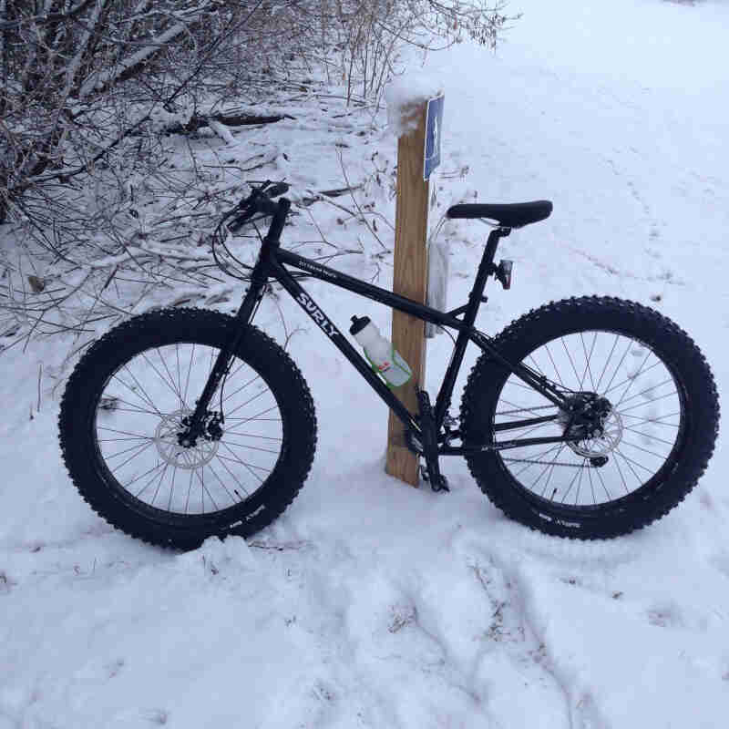 Left side view of a black Surly Ice Cream Truck fat bike, parked in the snow against a trail sign post in the woods