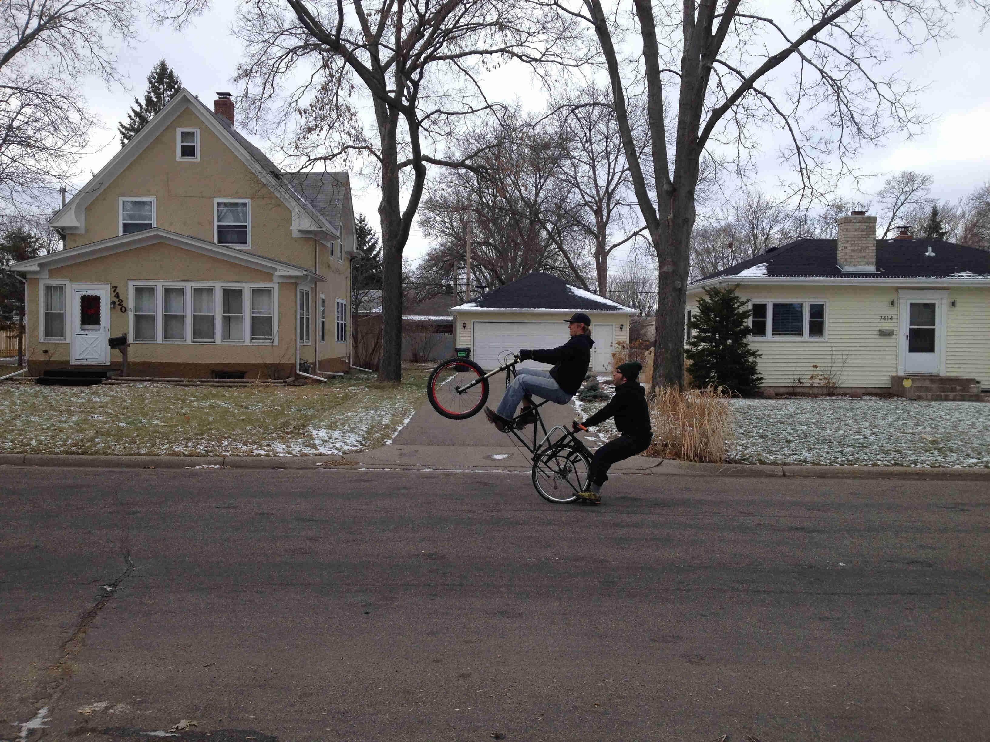Left side view of a cyclist riding a wheelie on a black Surly Big Dummy bike with a person on back, on a paved street