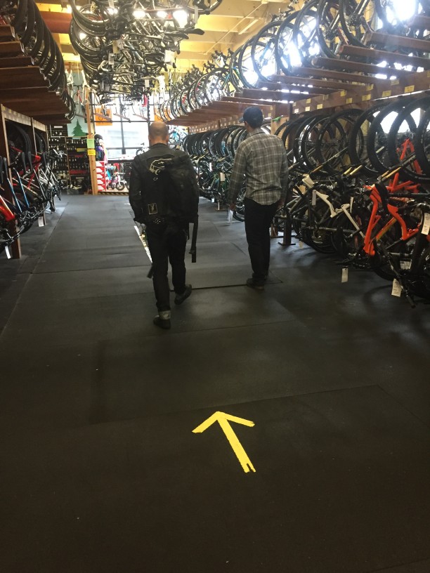 Rear view of 2 people walking between rows of a bikes, at a bike shop