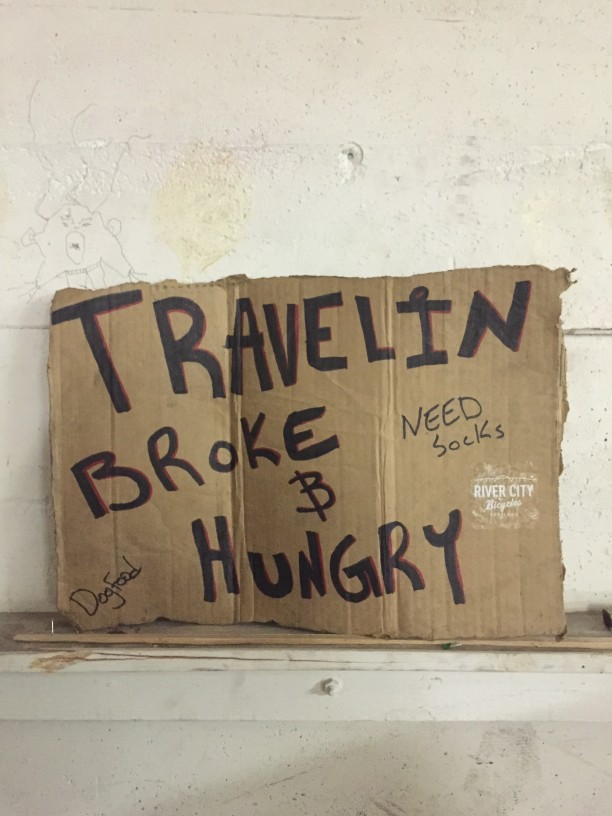 A cardboard sign against a cement wall that reads, Travelin Broke & Hungry