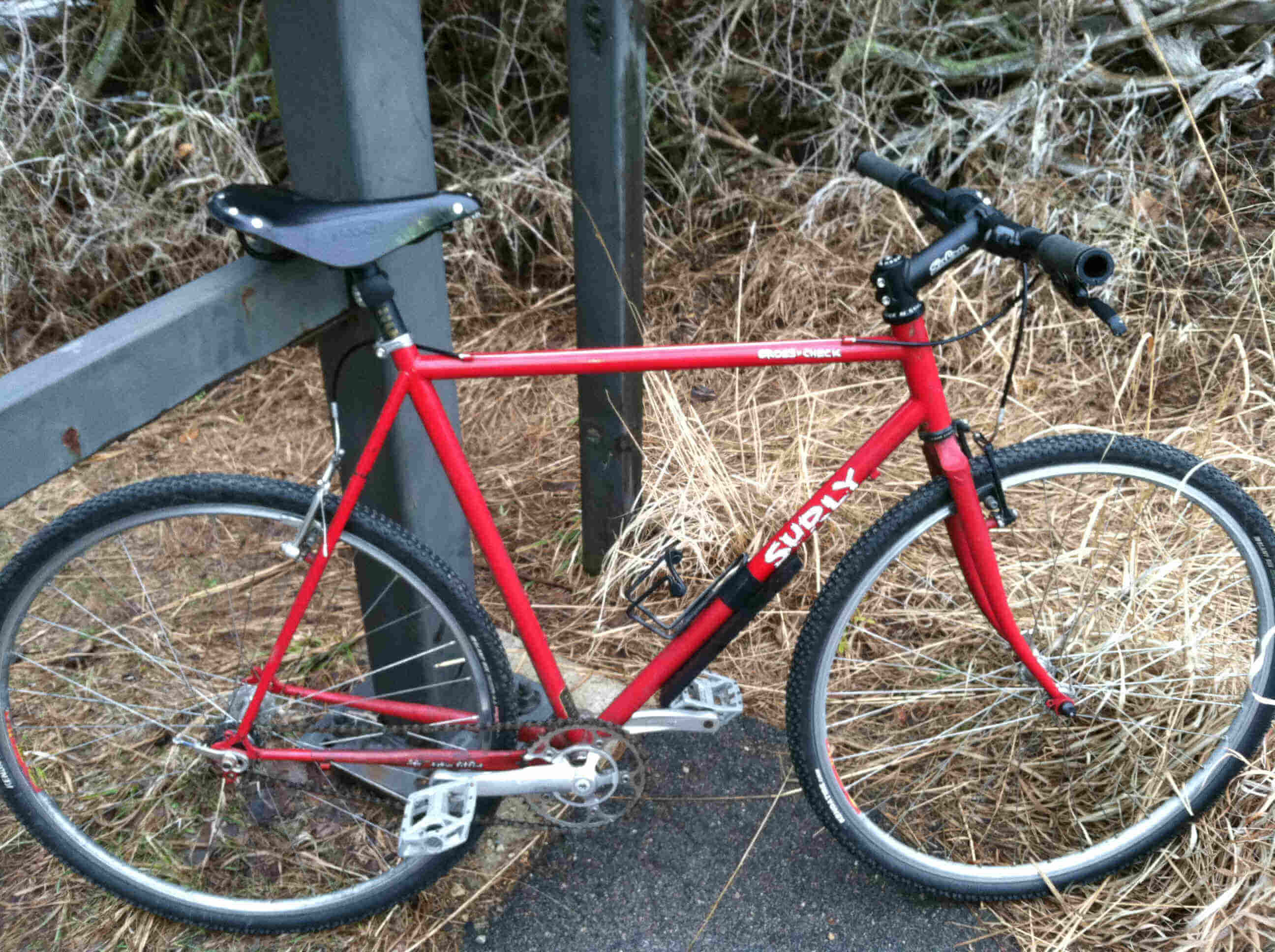 Right side view of a red Surly Cross Check bike, leaning on a steel gate post, with tall brown grass in the background