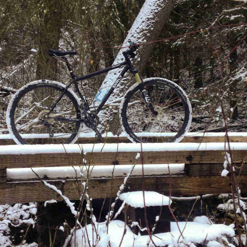 Right side view of a black Surly bike, parked on a wood crossover bridge in the snowy woods