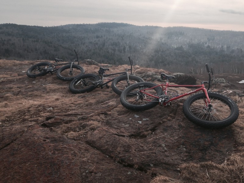 Right side view of 3 Surly fat bikes, laying on their sides at the edge of a rock cliff, with tree covered hills below