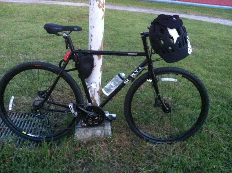 Right side view of a black Surly bike with a helmet on the handlebar, parked against a post in a grass field