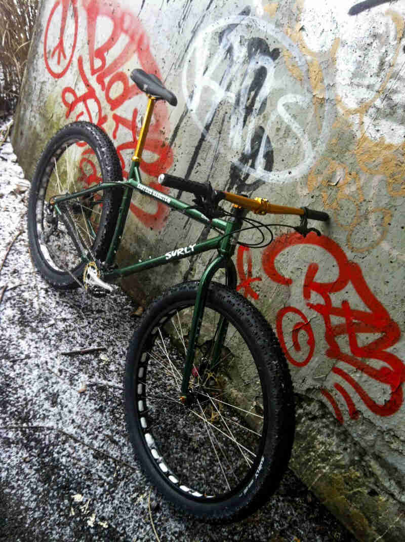 Tilted downward view of a Surly Krampus bike, green, leaning on a cement wall with graffiti