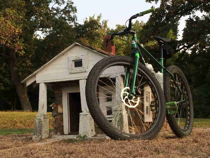 Front, ground level view of a Surly Krampus bike, green, in front of an aged log cabin, with trees in the background