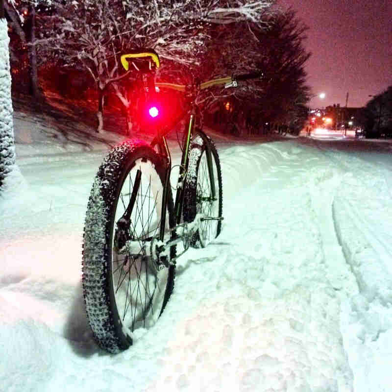 Rear view of a green Surly fat bike with a lit, red seat post light, parked in snow on the side of a street at night