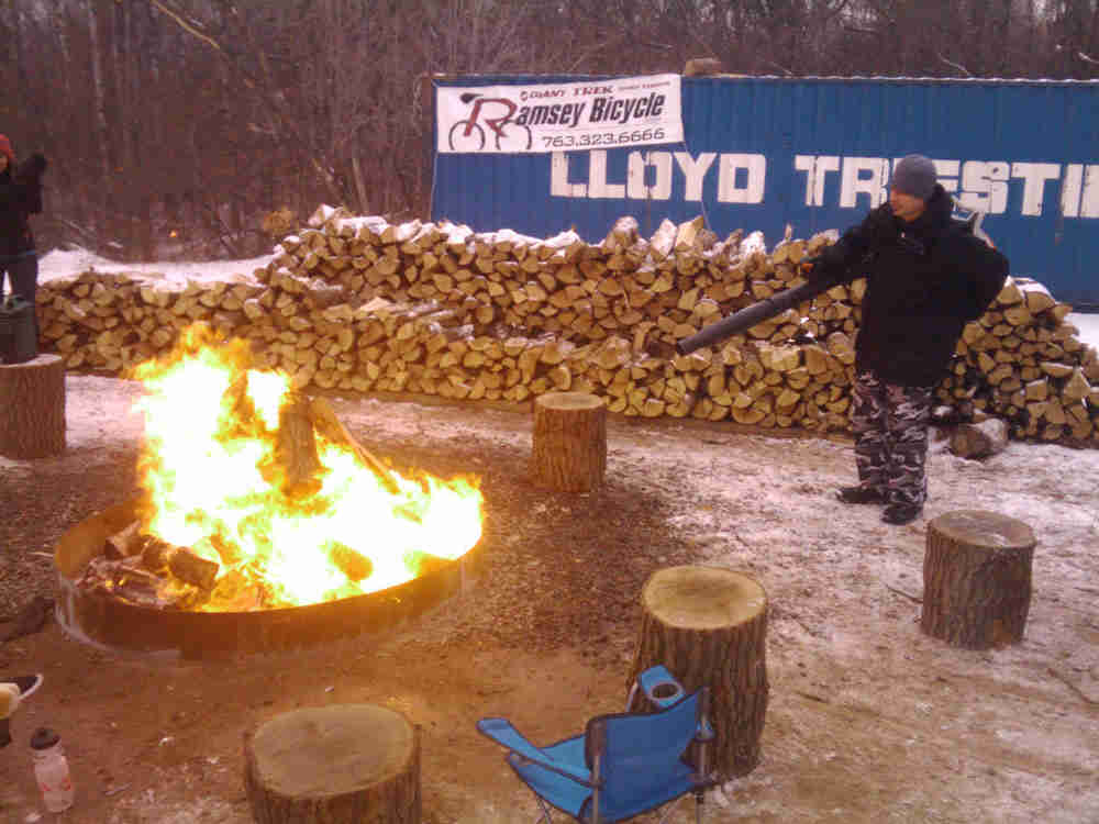 A campfire burning in snowy lot, with a person standing on the right, with a stack of split wood in the background
