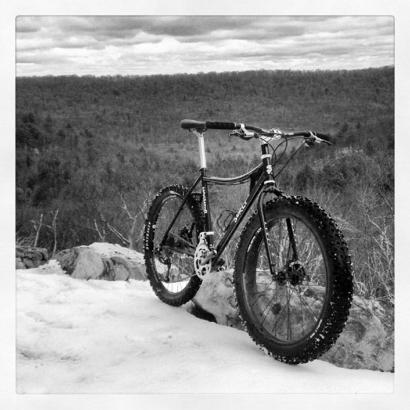 Front right side view of a Surly bike, standing on a snowy ledge, with a hilly forest in the background down below