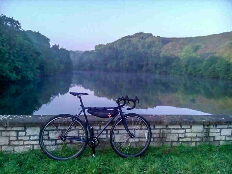 Right side view of a black Surly Cross Check bike, park in grass against a stone wall, in front of a pond