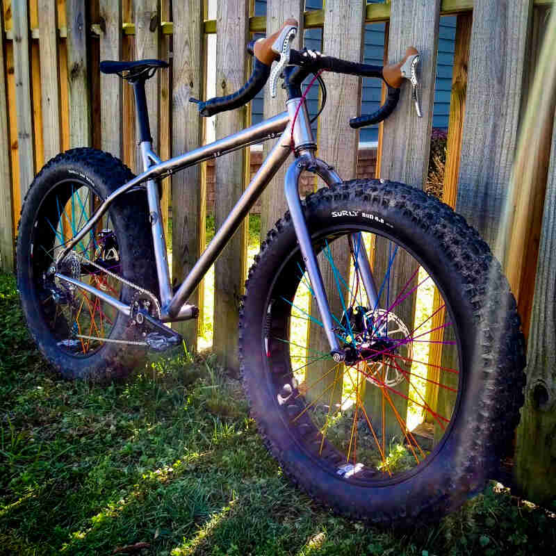 Front, right side, angled view of a silver Surly fat bike, on grass, leaning against a wood fence