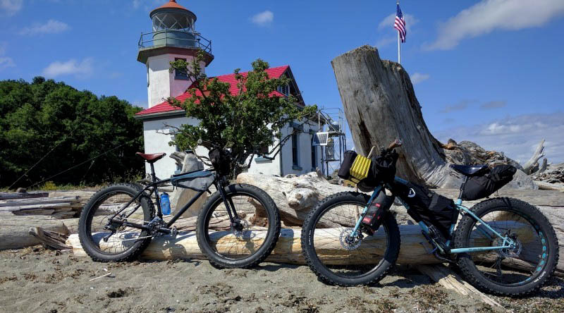 Side view of 2 Surly bikes facing each other, parked in sand with logs, with a lighthouse building in the background