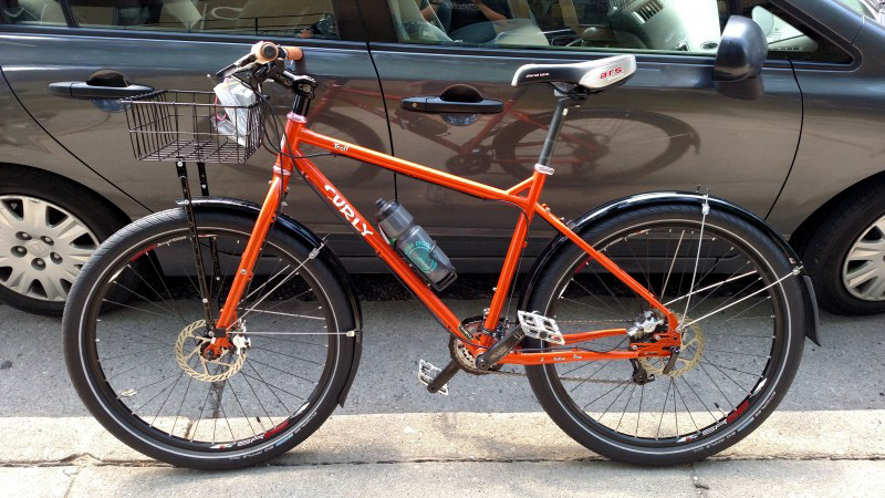 Left profile of an orange Surly Troll bike on a sidewalk, with a car on the street in the background