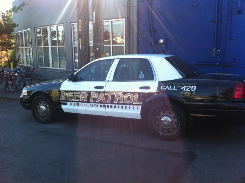 Left side view of a car, painted like a police car, with BEER PATROL painted on the side, parked next to a street curb