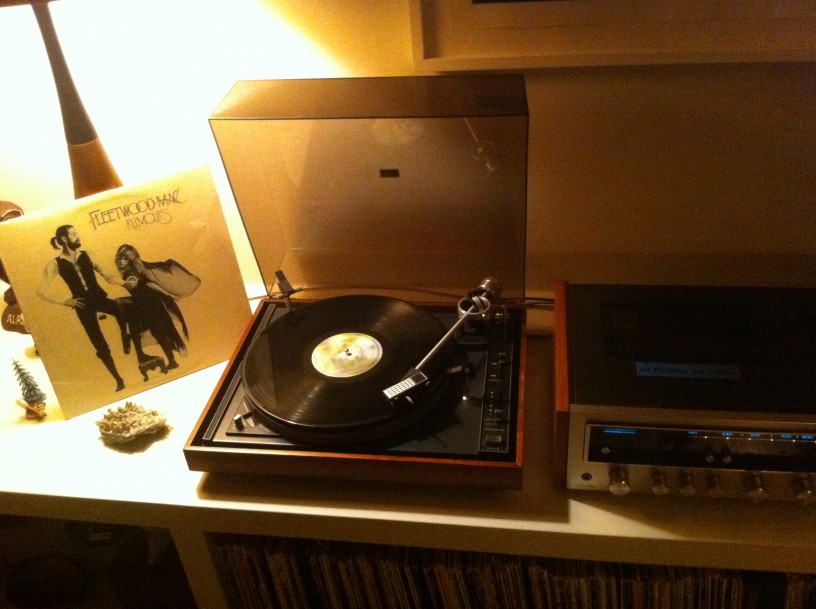 Downward view of a record player, a Fleetwood Mac album and a stereo tuner, on a cabinet top, against a wall