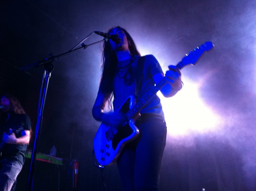 An upward, front view of a musician singing an playing a guitar, on a dark stage