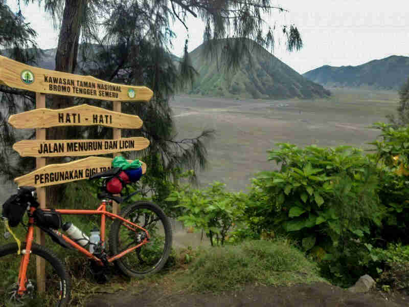 Left side view of an orange Surly Troll bike, leaning on sign, with a grass field and green mountains in the background