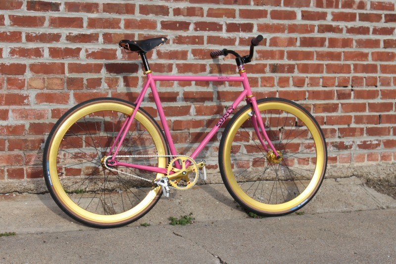 Right side view of a pink Surly bike with gold rims and cranks, parked on a sidewalk, leaning against a brick wall