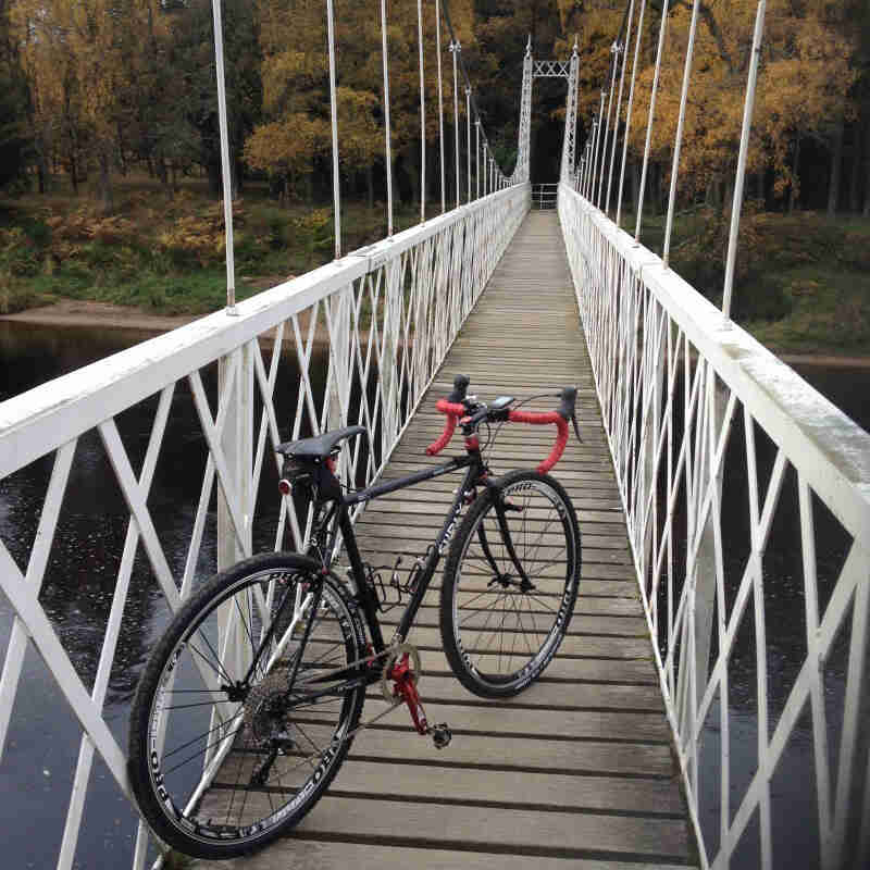 Rear right view of a black Surly Cross Check bike, parked on a trail bridge above a river, facing into the forest