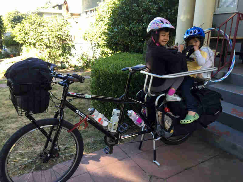 Left view of a Surly Big Dummy bike, black, with 2 small children in the rear rack, in front of a home