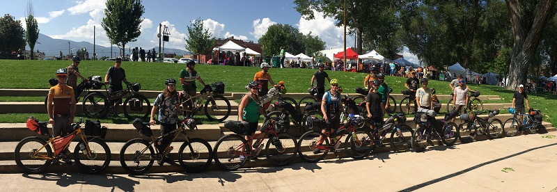 A group of cyclists standing with their bikes on a stretch of concrete steps, in front of a grass field with canopies