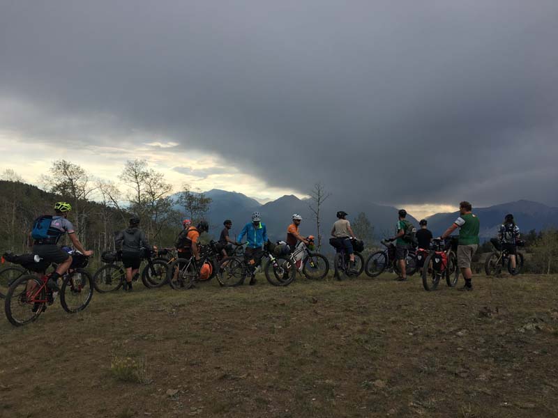 Cyclists lined up on a grassy plot, stare up at storm clouds above the mountains in the distance