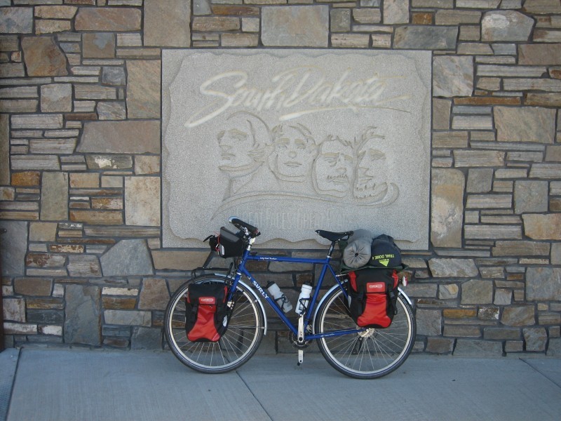 Left side view of a blue Surly Long Haul Trucker bike, loaded with gear, against a stone wall with a Mt. Rushmore sign