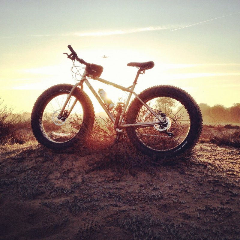Left side view of a Surly fat bike, parked on a dirt mound, with the sun behind on the horizon and a plane flying over
