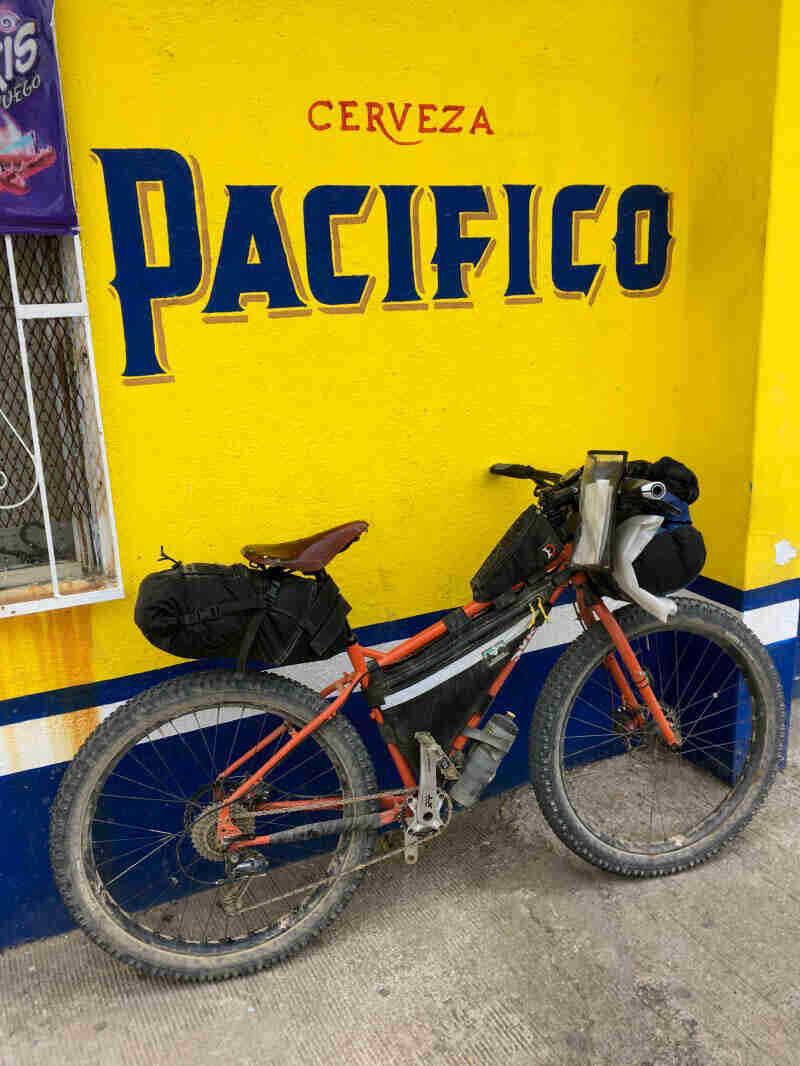 Right side view of an orange Surly bike, loaded with gear, leaning on a yellow wall with Cerveza Pacifico painted on it