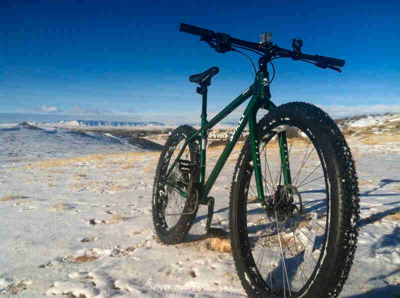 Front, right side view of a Surly bike, green, on snowy plains, with mountain in the background and blue sky above