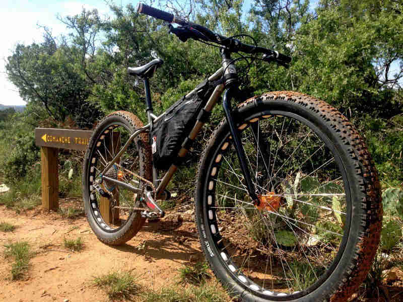 Front right side view of a Surly Krampus bike, parked on desert trail, in front of small cactuses and trees