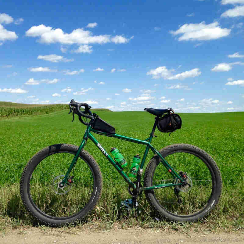 Left side view an emerald green Surly bike, parked on the edge of a grass field, next to a cornfield