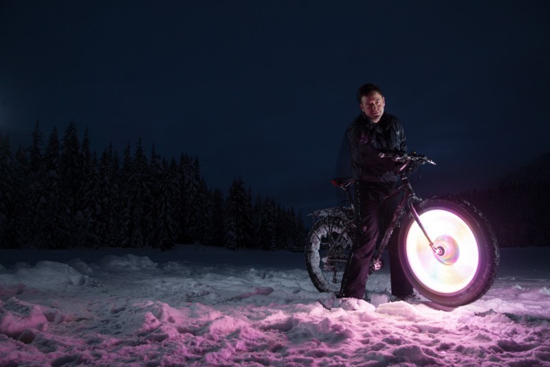 Right side view of a Surly fat bike, with a lit up front wheel, and cyclist standing over, on a snowy field at night