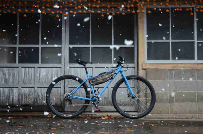 Right profile view of a blue Surly bike on pavement, in front of a building with windows, with snow falling