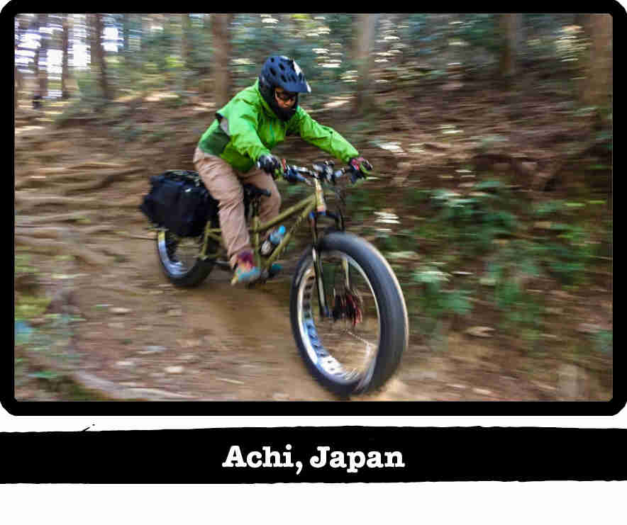 Right side view of a cyclist riding a Big Fat Dummy bike down a wooded, dirt trail hill - Achi, Japan tag below image