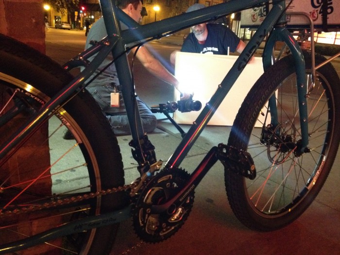 Low, right side view of a blue Surly bike, parked on a sidewalk, with 2 people kneeling with a light & camera, at night