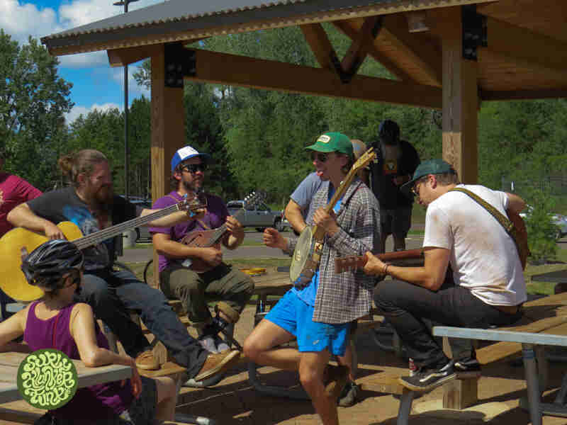 A group of musicians playing instruments, while sitting on picnic tables, under a park shelter