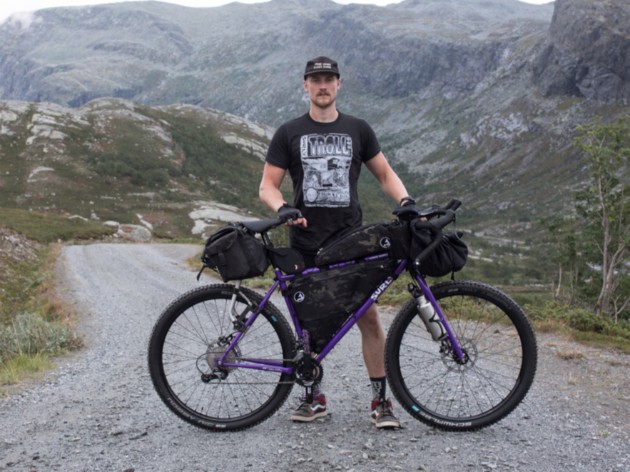 Cyclist poses while standing with a purple Surly bike on a gravel road in the mountains