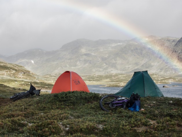Two tents and two bike laying on a rocky grass clearing in the mountains with a rainbow arching over